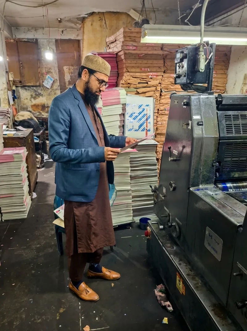 Muhammad Yaseen checking a freshly printed sheet beside an offset press